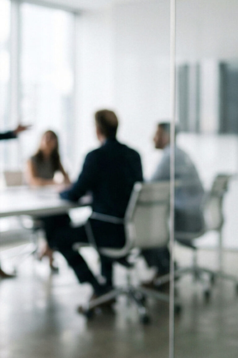 A business meeting takes place behind a glass door, showcasing professionals discussing strategies in a modern office setting with a focus on teamwork.